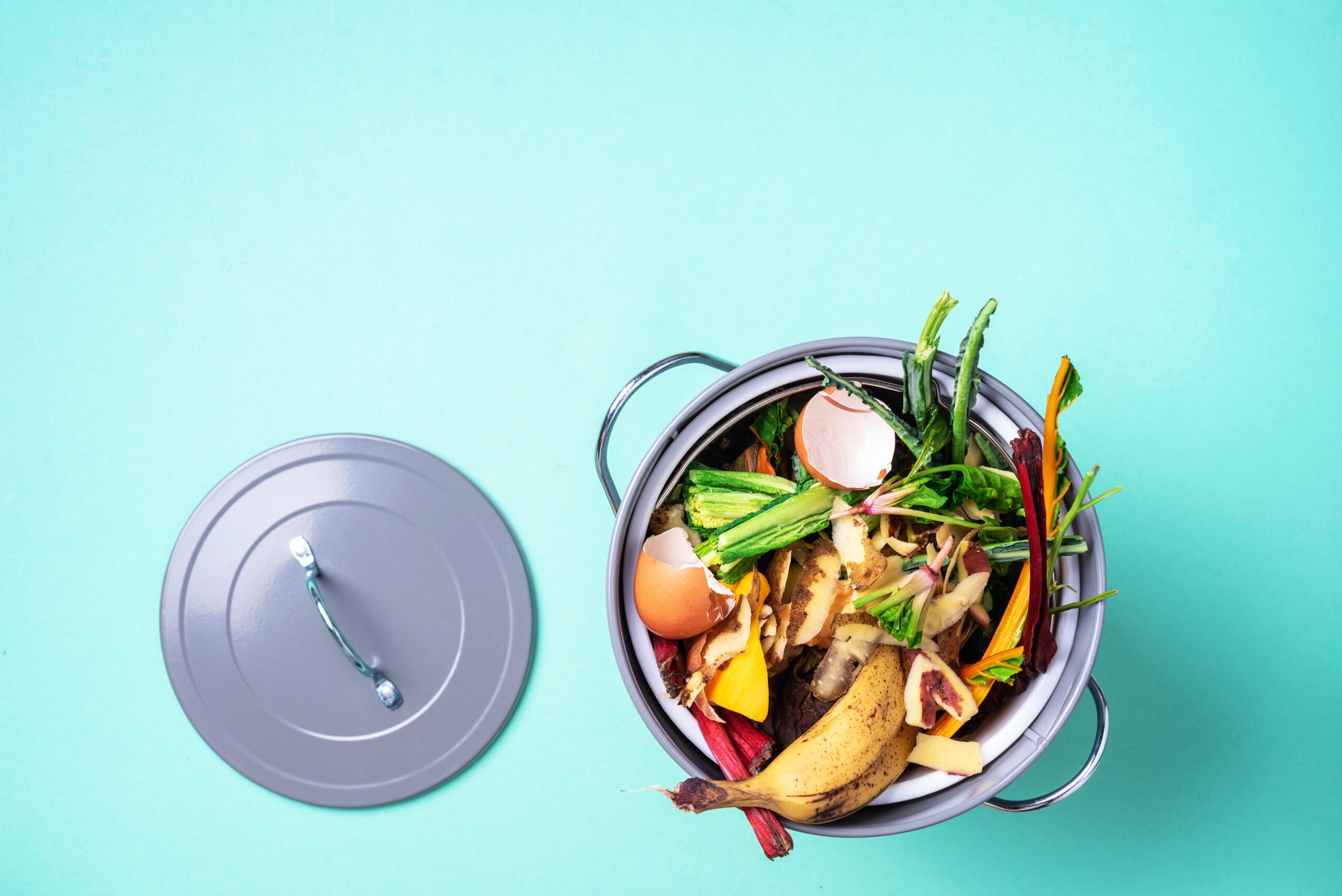 Peeled vegetables on chopping board, white compost bin on blue background. Top view of kitchen food waste collected in recycling compost pot.